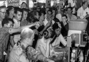 College-aged students (white male, white female, and black female) sitting in at Woolworth lunch counter performing non-violent direct action are being heckled by a group of college-aged and older white males. One white male is seen actively dumping food items on the head of the white female activist. The heads of the other activist (white male and black female) are both covered with food items such as ice cream, drinks, and sugar that were dumped on them.