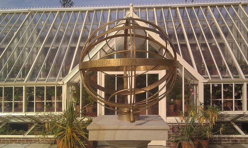 In front of a greenhouse, large bronze rings fit inside each other in a spherical shape.