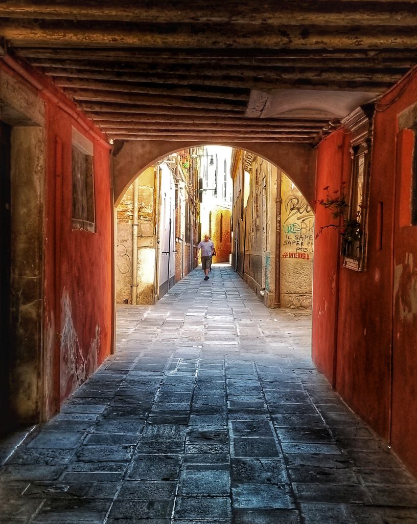 Photograph of a back street in Venice, Italy with red architecture and a blue cobblestone street