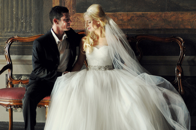 A man in a tux sitting on a bench with his newly made wife (in her wedding dress)