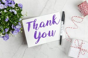 Small bunch of violets, thank you note in purple ink, with a pen beside it, and two small presents, all displayed on a white surface.