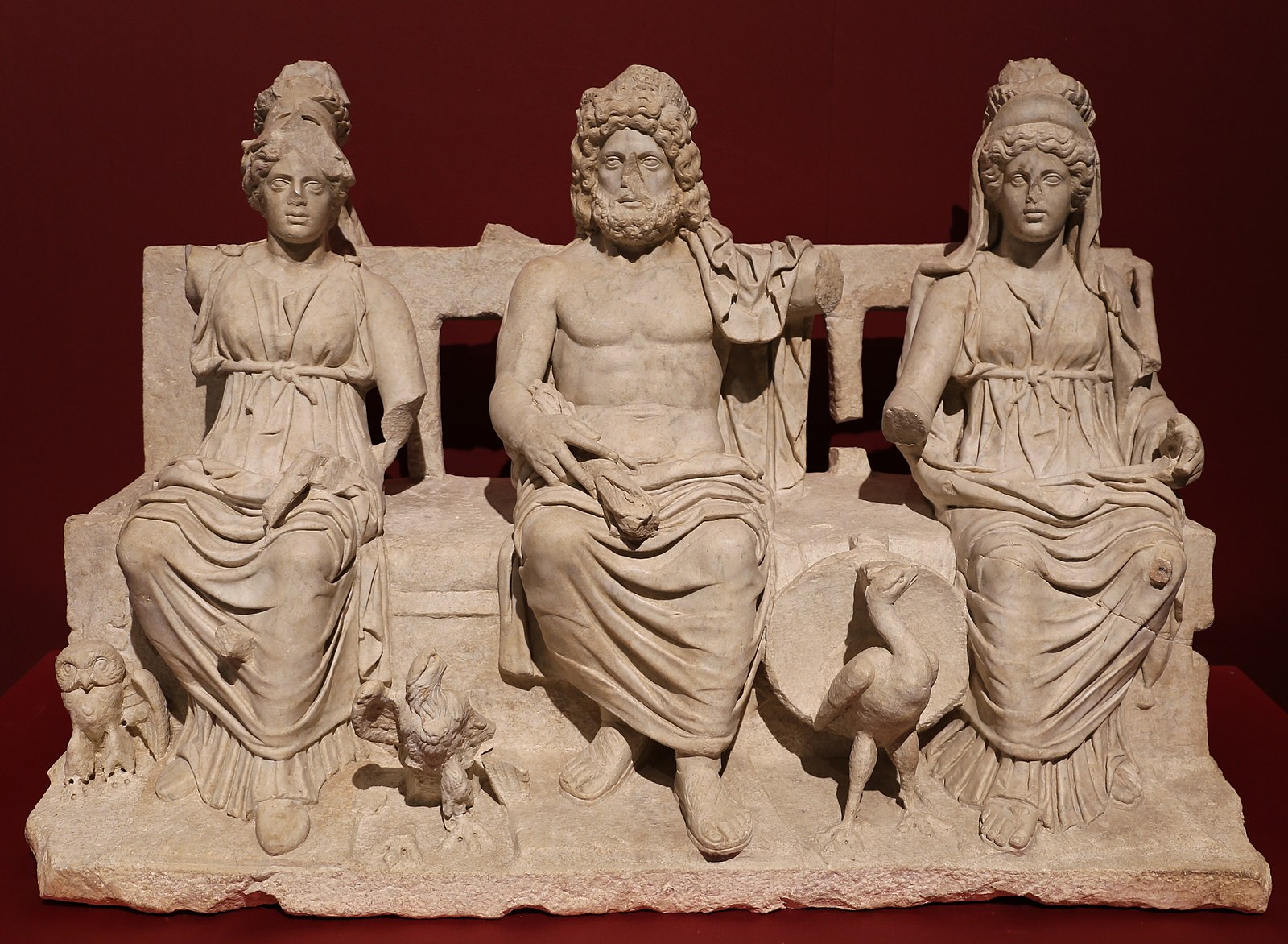 Sculpture of three ancient Roman figures (two women with a man in between) seated on a bench, wearing togas and formal headdresses. Missing some limbs and noses are chipped off.