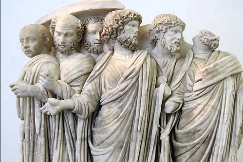 Marble statue of six male figures standing in a line (one head is missing), wearing togas and wiht different body positions and expressions