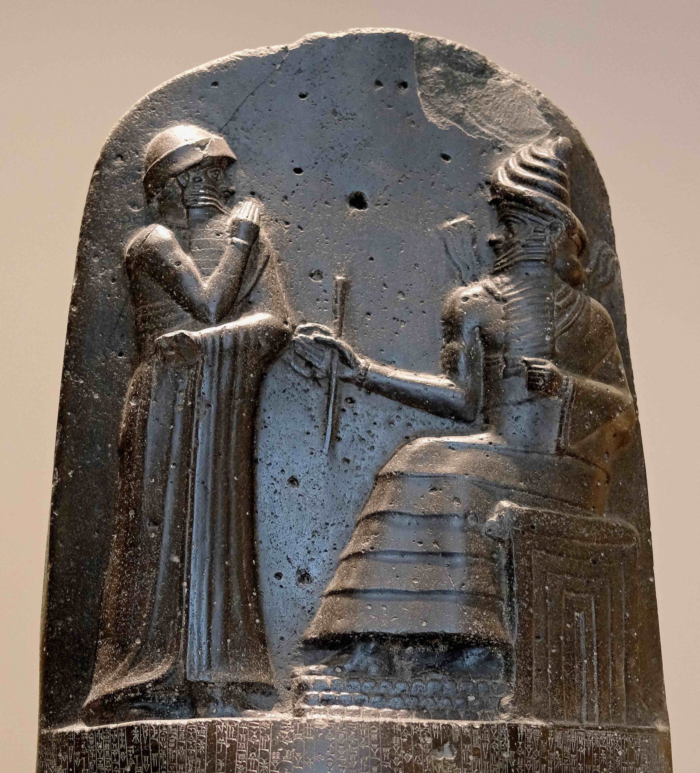 Stone image of man standing with hands in front before seated man on throne.