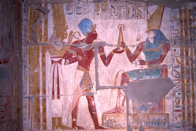 Painted image showing three figures with elaborate headresses and ancient Egyptian clothing. One is receiving a gift from a figure with a hawk head.