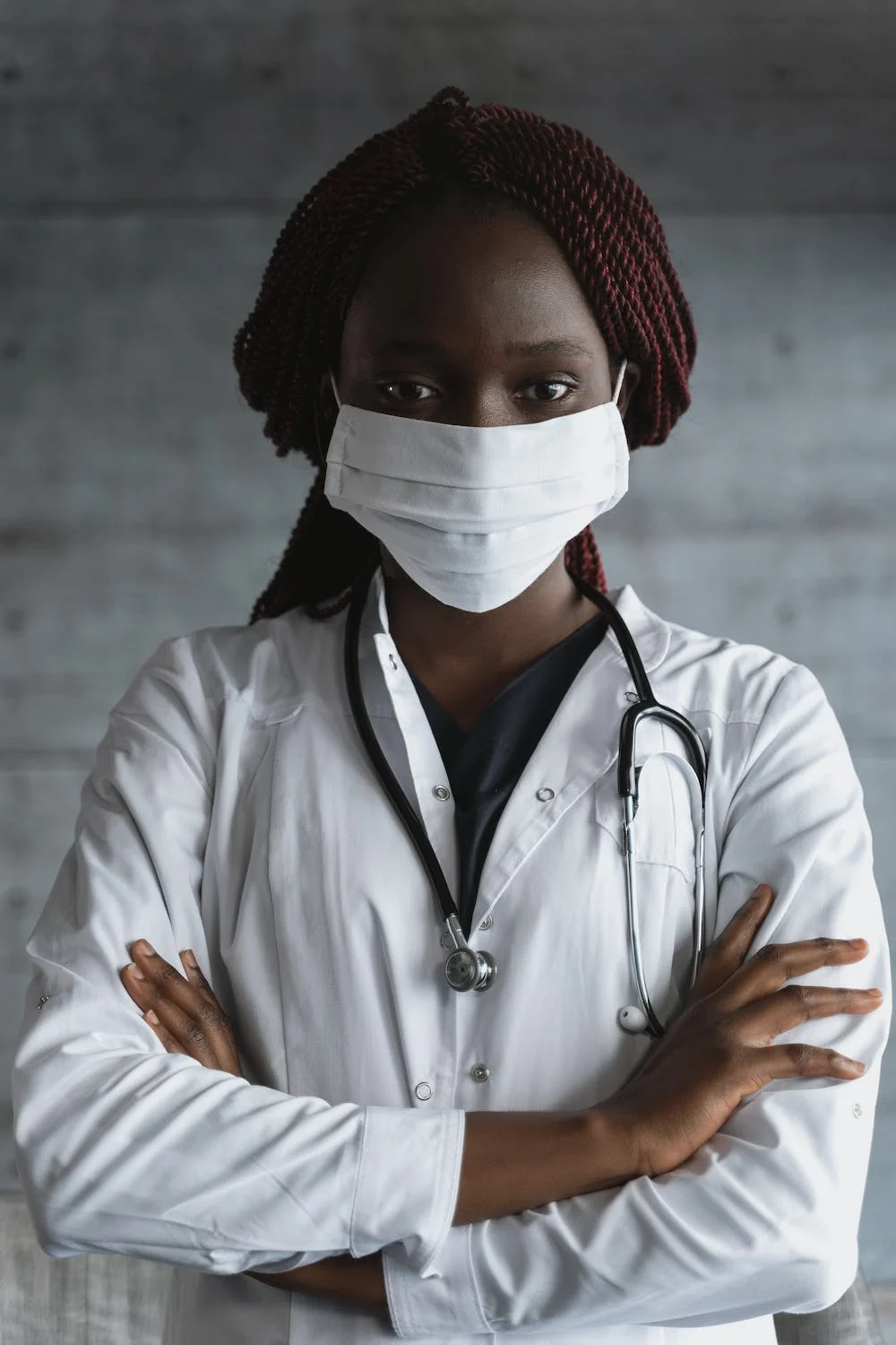 A young black woman in a white face mask is pictured from the waist up, wearing a white lab coat and has a stethoscope around her neck Her arms are crossed.