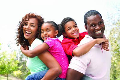 Black woman is smiling and carrying a smiling Black girl on her back, posed beside, but turned away from a smiling Black man carrying a smiling Black boy on his back. All are dressed in casual clothes.