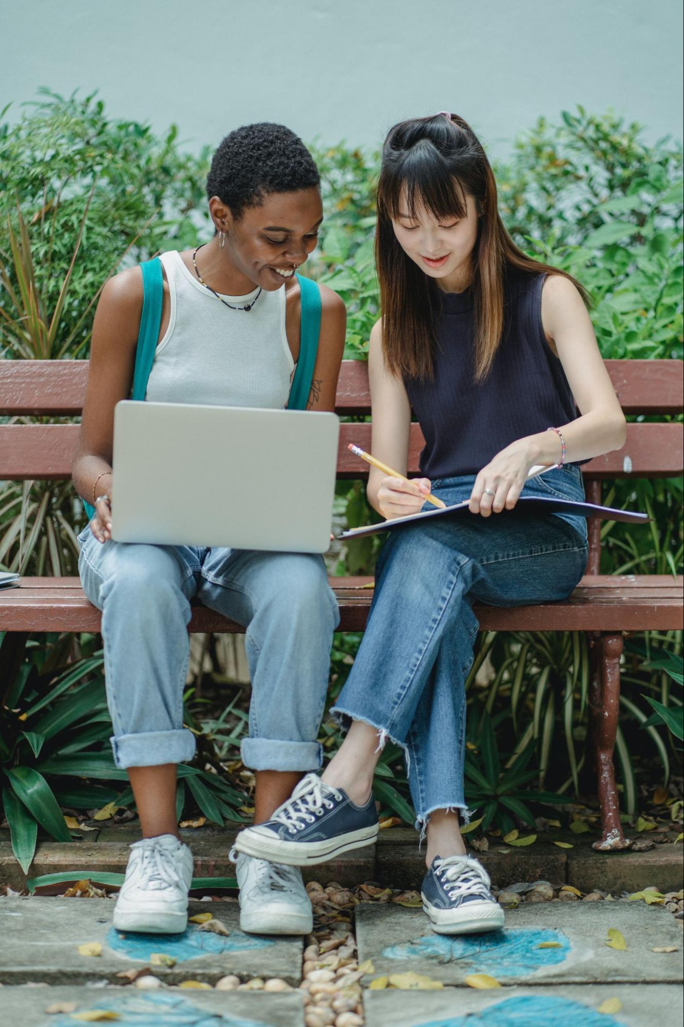 Two students sit on a bench and study together with one holding a laptop and the other writing in a notebook.