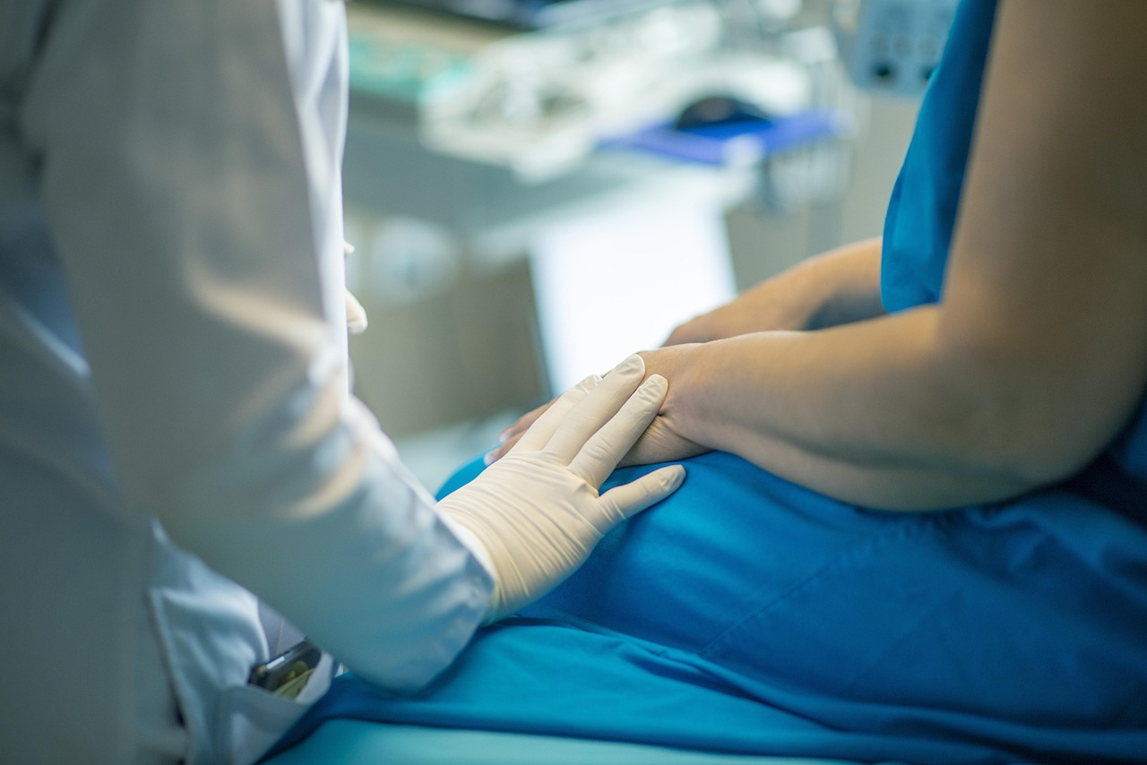 A person with a white coat and a glove on their hand is resting their fingers on the hand of a patient in a blue gown. Only the arms are shown.