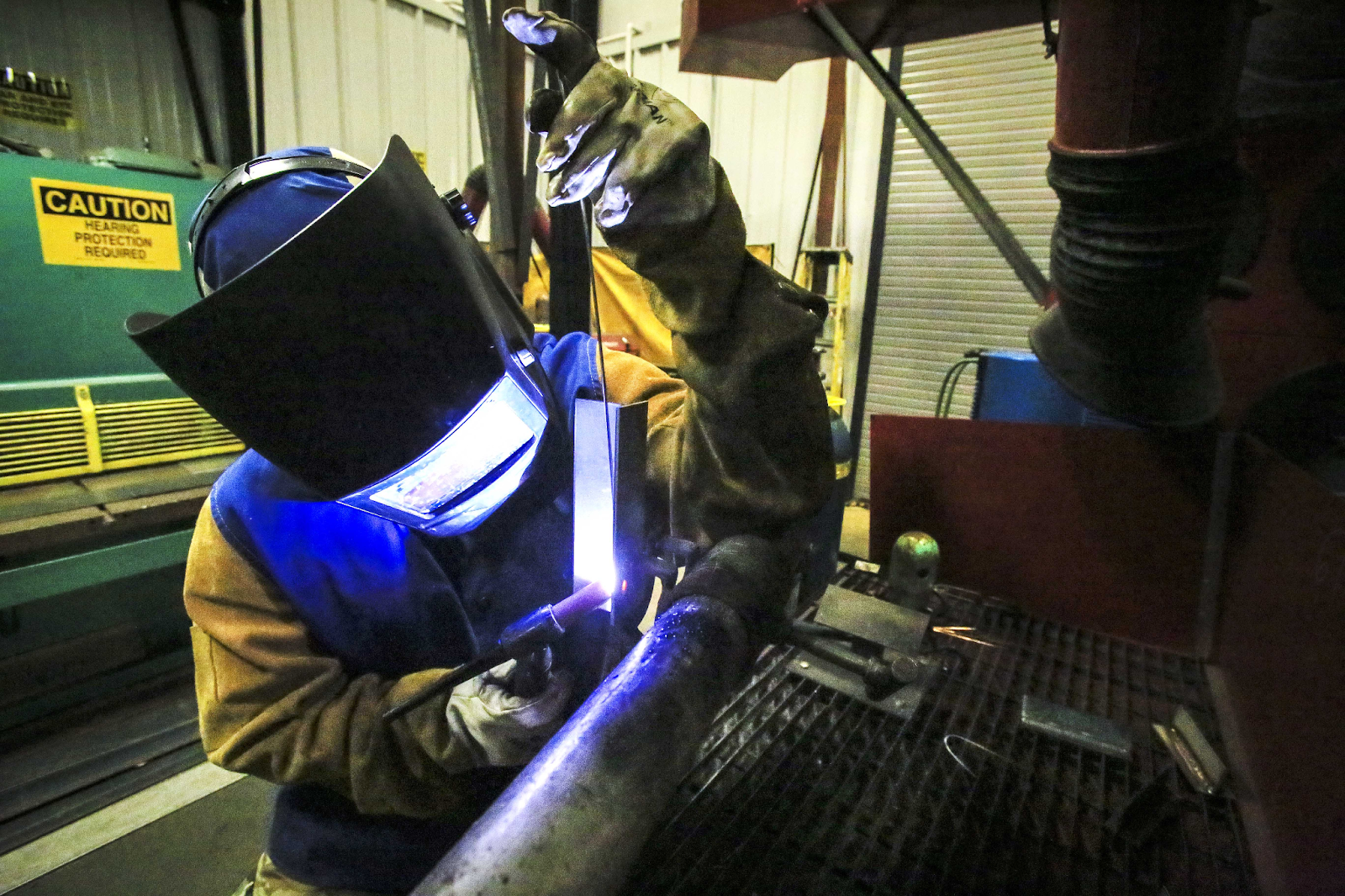 Person wearing a protective welding jacket and welding hood, welding a practice piece vertically using GTAW