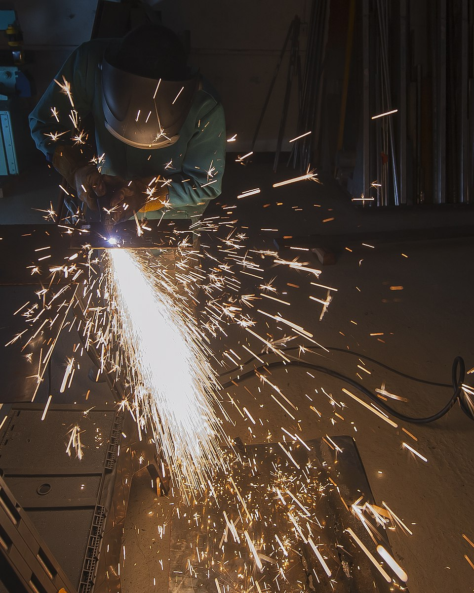 A person wearing safety gear, including a helmet with a faceguard performs plasma cutting. Sparks generated by the cutting process flash from the metal being cut.