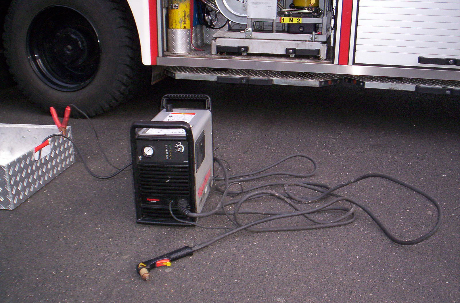 A plasma power source is hooked up to an aluminum box for grounding. The plasma power source is a rectangular shaped box with dials on the frog and slots for cables to be plugged in.