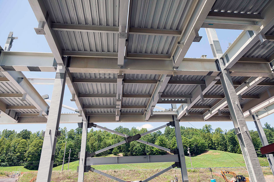 inside a steel building. Steel supports are welded together and a steel roof is pictured. The building doesn’t have finished walls, so the green of the hillside in the background can be seen.