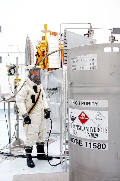 Stainless steel hydrazine tank inside of a white laboratory room. The tank is a cylinder with a hose leading away from it.