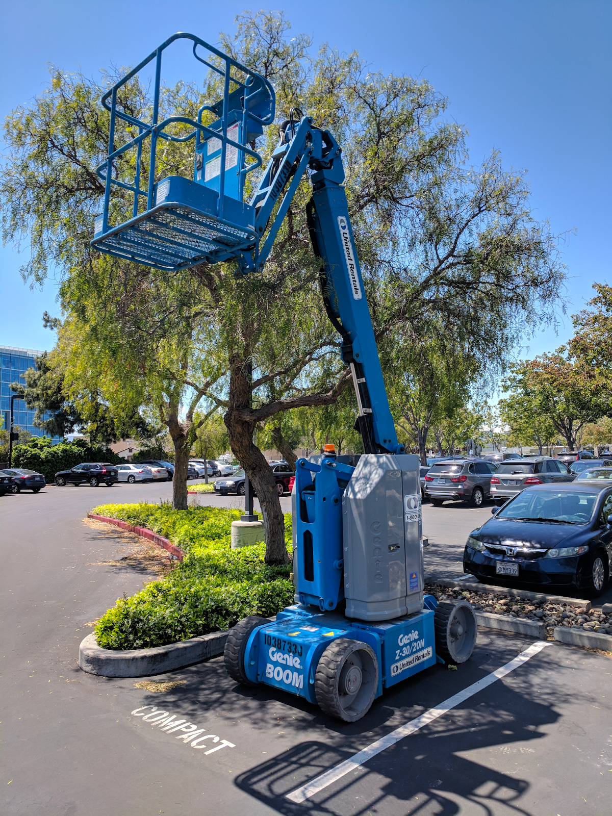 A small blue lift with an articulating arm is parked outside in a parking lot next to a car.