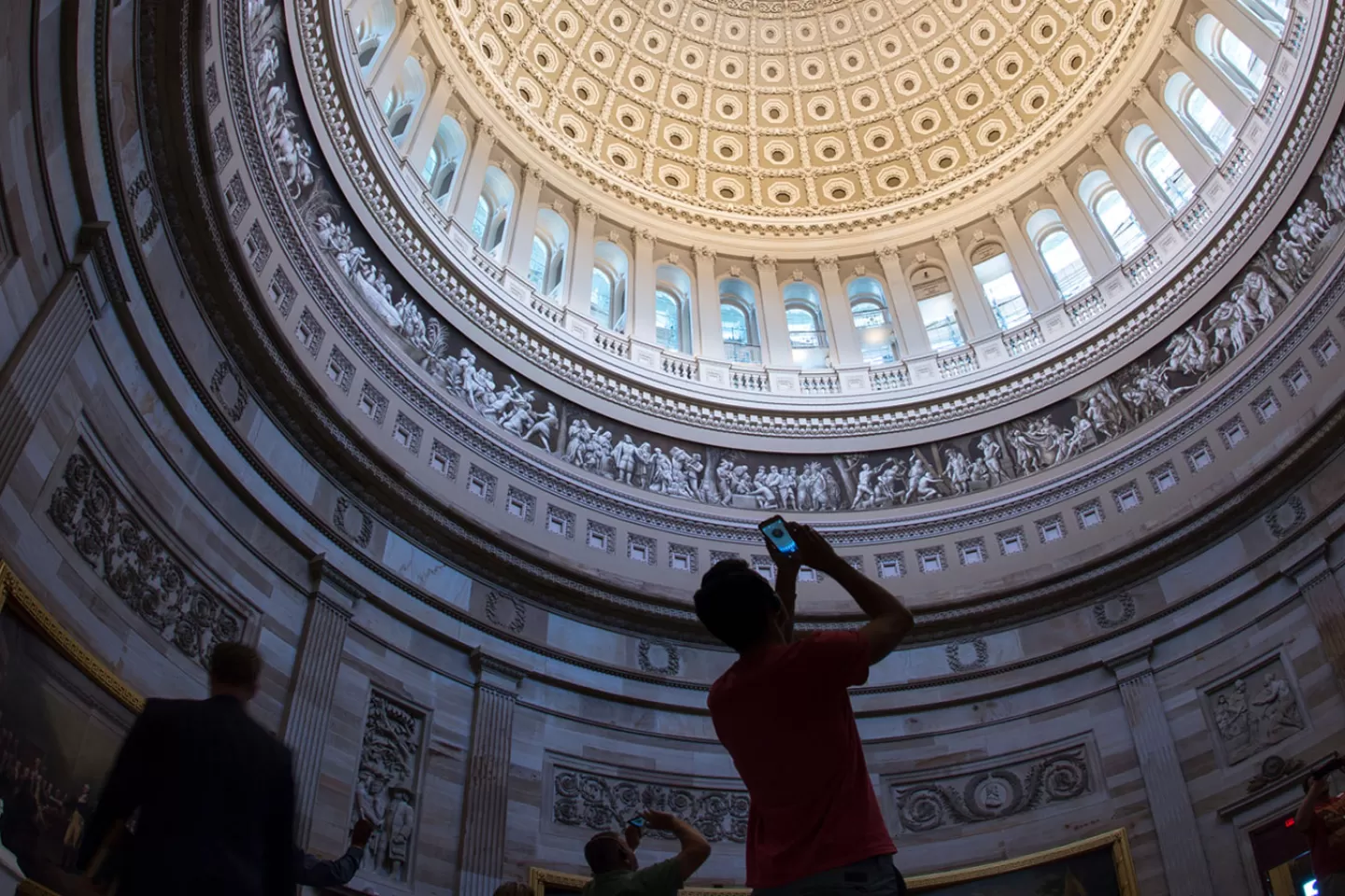 The US Capitol Dome Rotunda. People stand below the dome and take pictures of the interior of the dome.