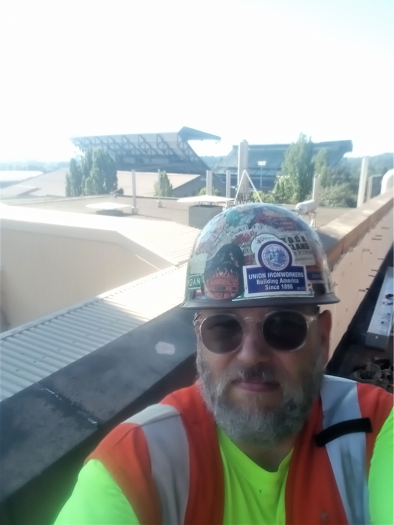 A man in a hardhat with dark glasses and a construction vest is on a rooftop outdoors.