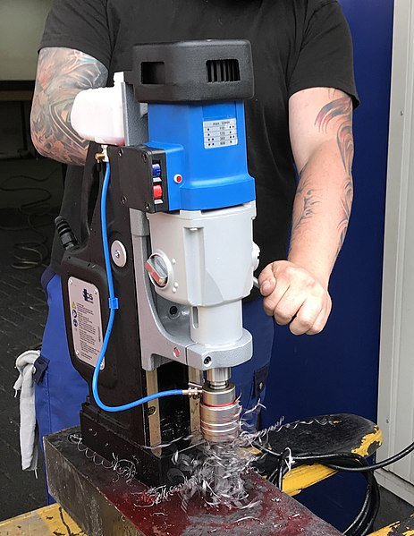 A person wearing a black shirt and blue pants holding a blue and gray magnetic drill on a piece of metal while operating the drill. The drill has a vertically mounted motor that slides up and down on a track, and is attached to a large vertical base with a magnet on the bottom. The spindle is mounted in line with the motor so that it is pointed downward. The operator is turning a handle on the back side of the machine which is forcing the drill down into the work piece.