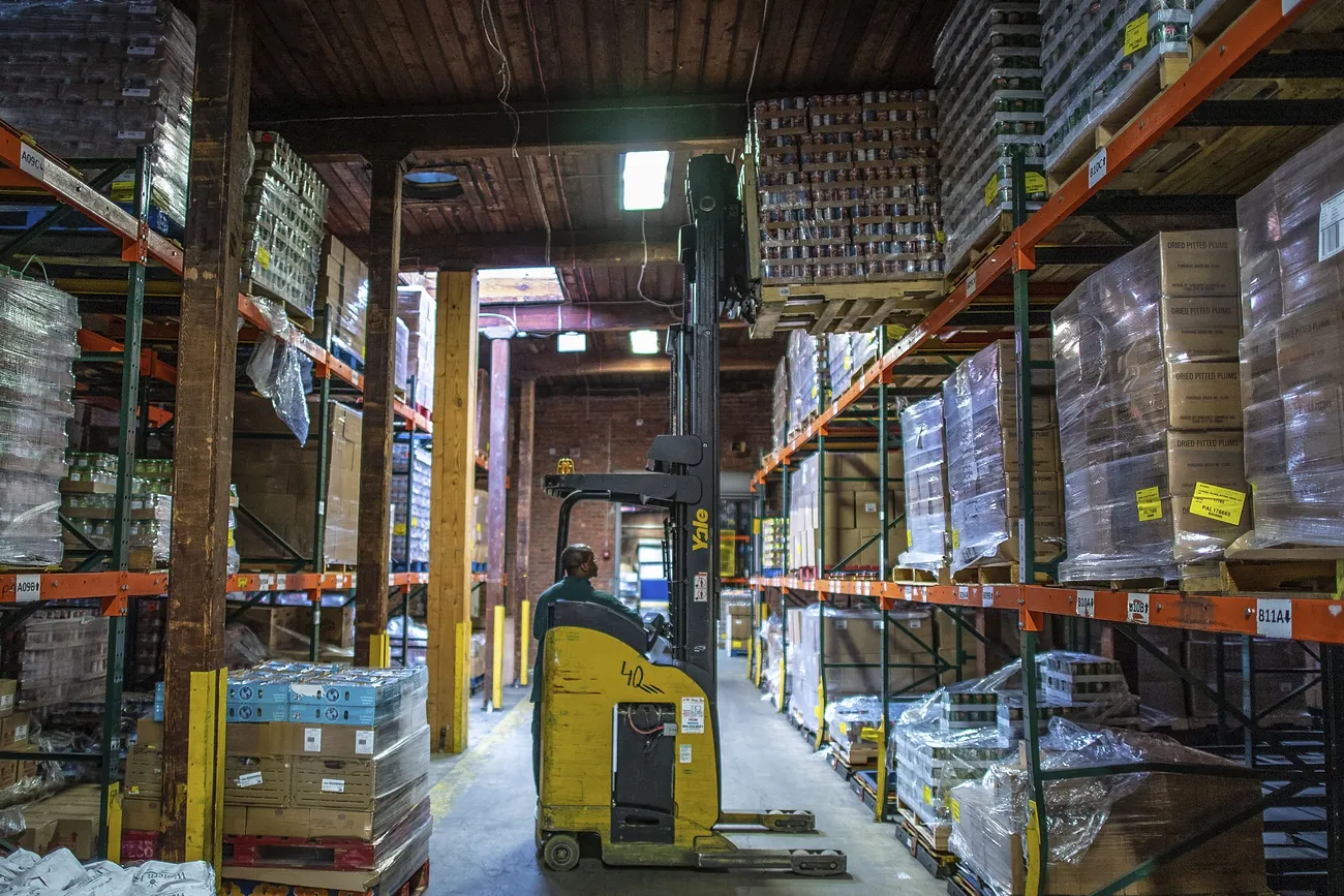 A forklift moves a pallet load of material to a shelf high overhead in a warehouse.