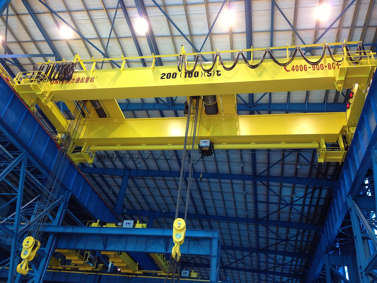 A yellow bridge crane is seen in a shop beneath a blue ceiling.