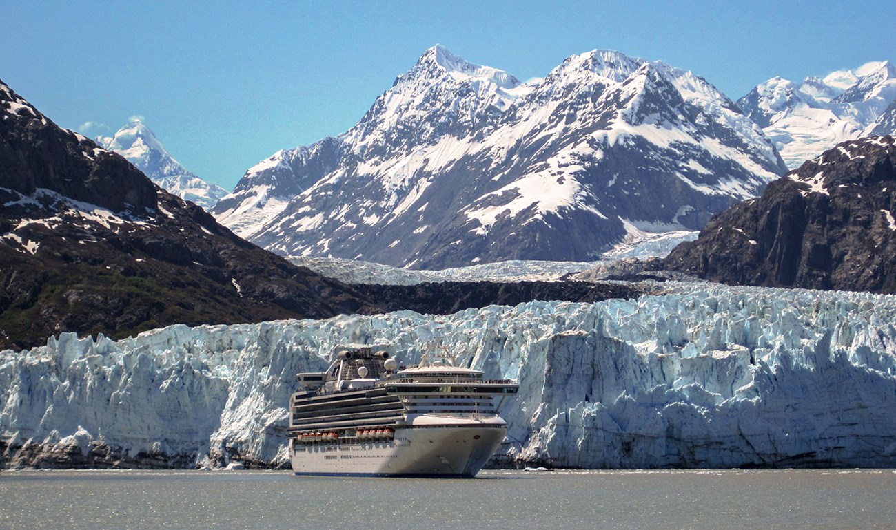 A photograph of a tan colored cruise ship as it sails in front of a snowy glacier with a dramatic mountain in the background.