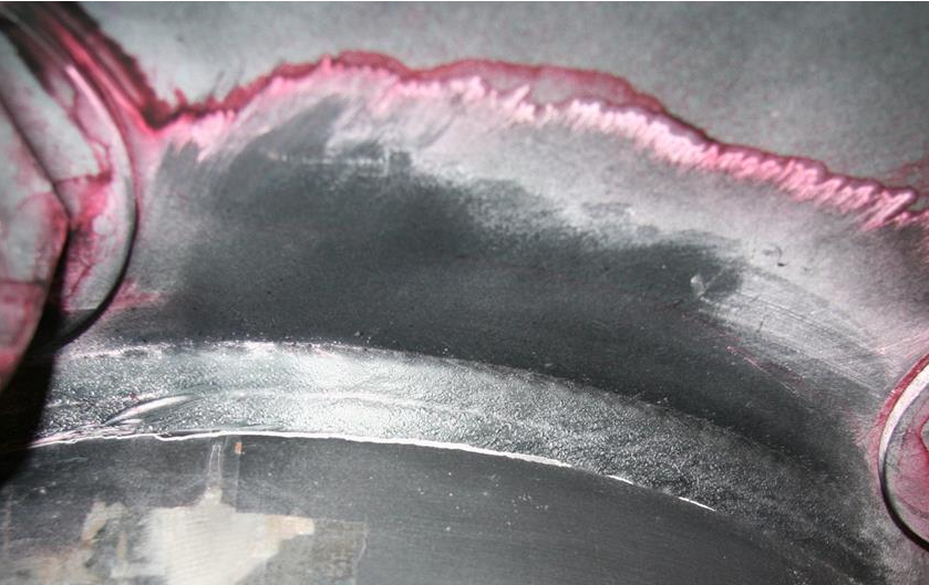 A close up of a weldment with an uneven red line about 2 inches above the weld bead and red surrounding two bolt holes. This is a crack seen on a sample that has been liquid penetrant tested. The crack is visible because the penetrant is red.