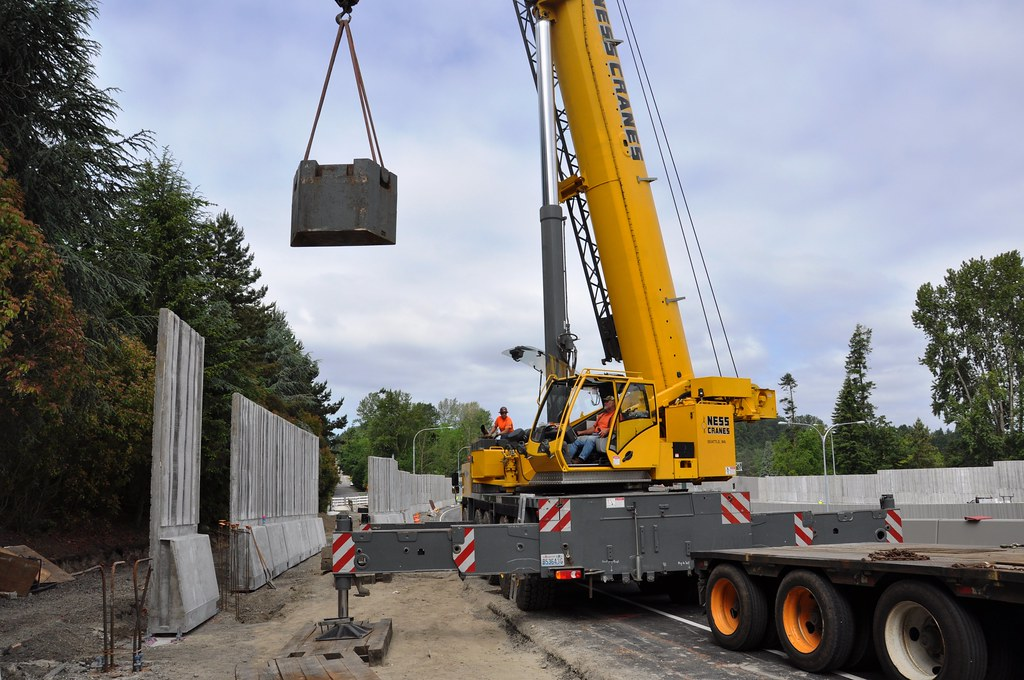 A ground crane with outriggers extended lifts a heavy counterweight at the side of a highway.