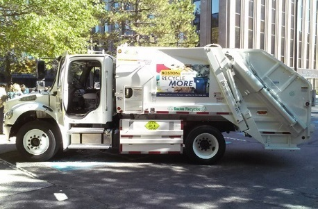 A side view of a white garbage truck with the driver’s side door open parked on a city street. Green trees and a tall building are in the background.