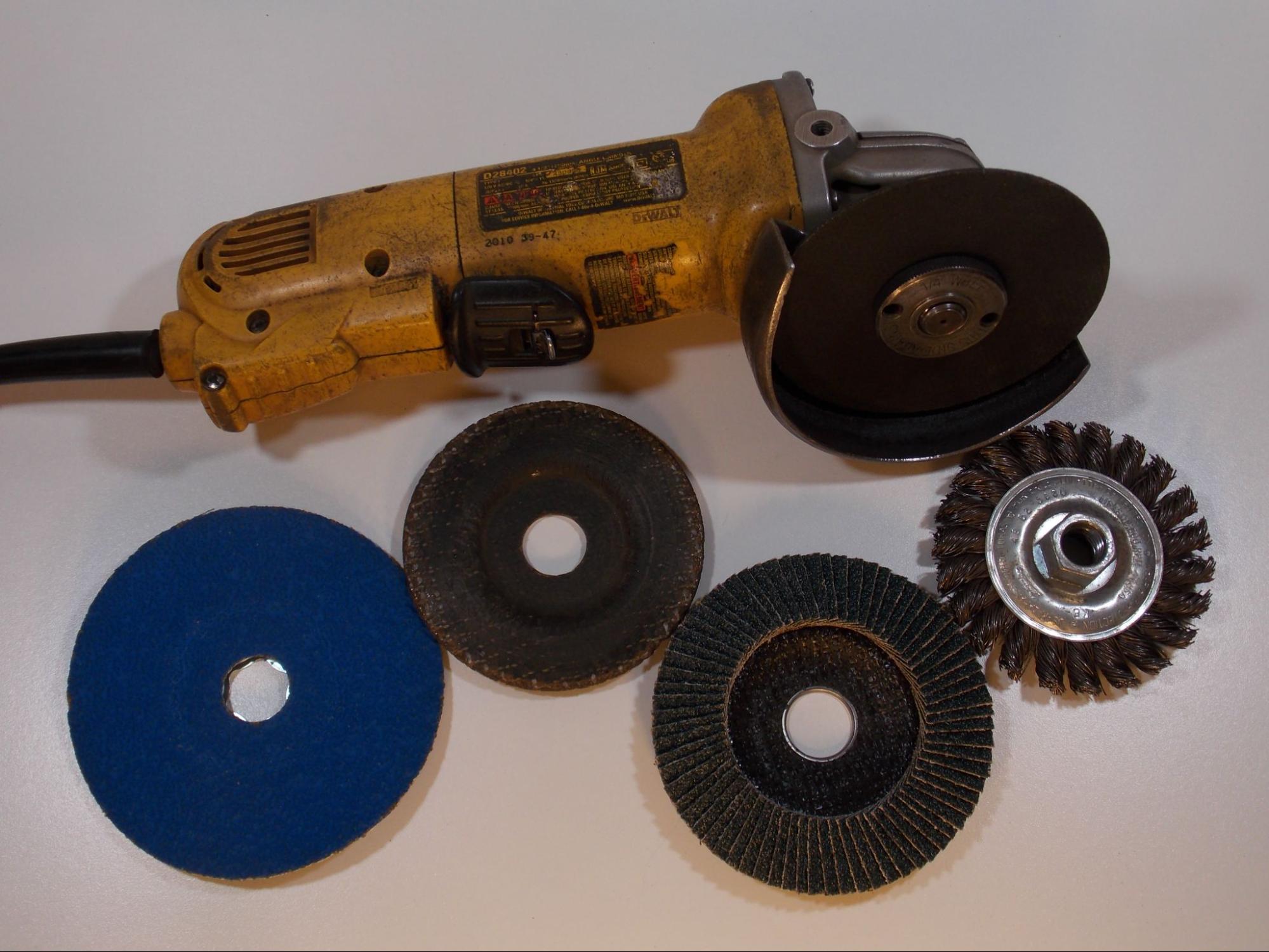 A common angle grinder. This grinder is yellow with a black power cord and paddle type switch. The tool has a threaded spindle at the head to which different abrasive discs can be mounted. The angle grinder is surrounded by four different examples of abrasive discs.