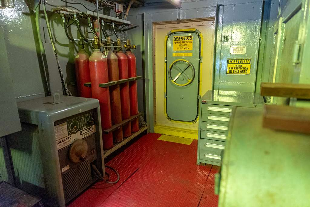 A large, gray transformer welding machine sits against the wall in a corridor aboard a ship. Next to it, several high pressure gas cylinders stand connected to a gas distribution manifold. At the end of the corridor is a large, gray toolbox and a closed bulkhead door with a wheel lock, which provides access to the next room.