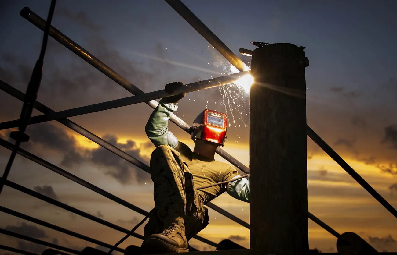 A welder welds outside, connecting what appears to be framing supports.