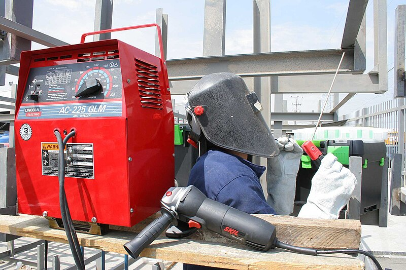 A welder uses SMAW to weld on steel beams. The welder is facing away from the red machine that is powering the arc being used on the steel beams.