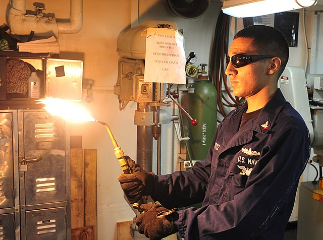 A sailor wearing protective goggles, gloves, and long sleeves adjusting the flame in his welding torch. The flame is currently at a carburizing flame.