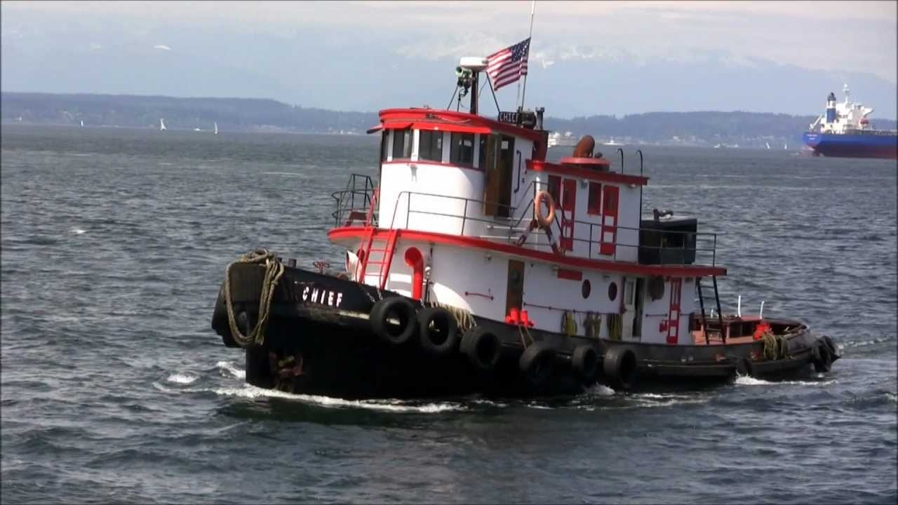 a tug boat with a black base, and a red and white cabin. The tug boat is in a body of water, likely the Puget Sound. In the background is a larger ship, probably used for shipping purposes and several sailboats.