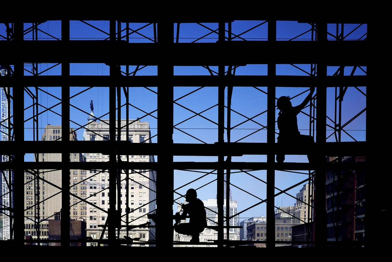 Two workers are silhouetted as they work on the steel frame of a building, with other highrises in the background.