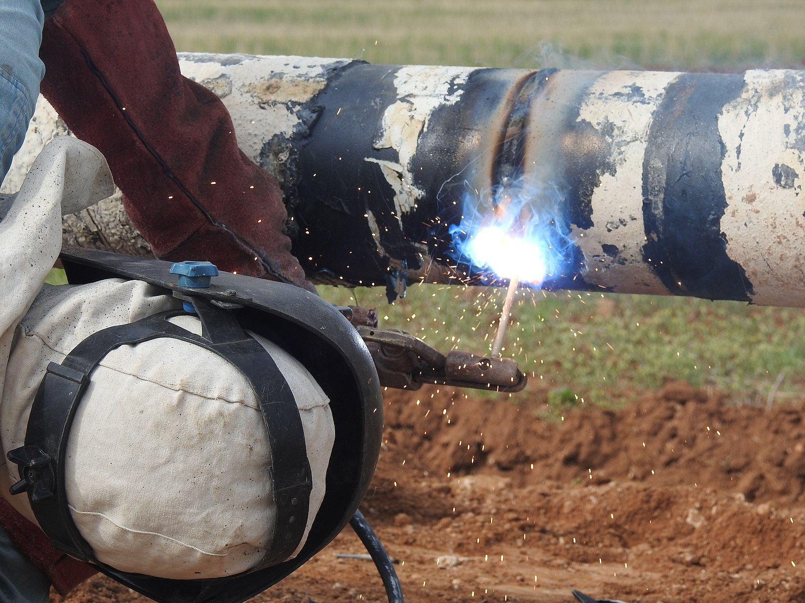 A welder lies on the ground while SMAW welding a pipe.