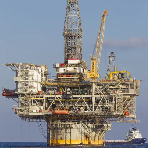 An offshore drilling platform is pictured. It has a large cylindrical base with an enormous scaffolding with cranes attached. A passenger ship is next to the platform on the right.