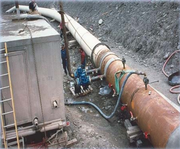 Leak Test of a pipeline showing a hose pressurizing the pipe. A worker sits on top a large box-shaped van or trailer and watches as a hose attached to the van and to the pipe operates the test. Another two workers stand on the ground.