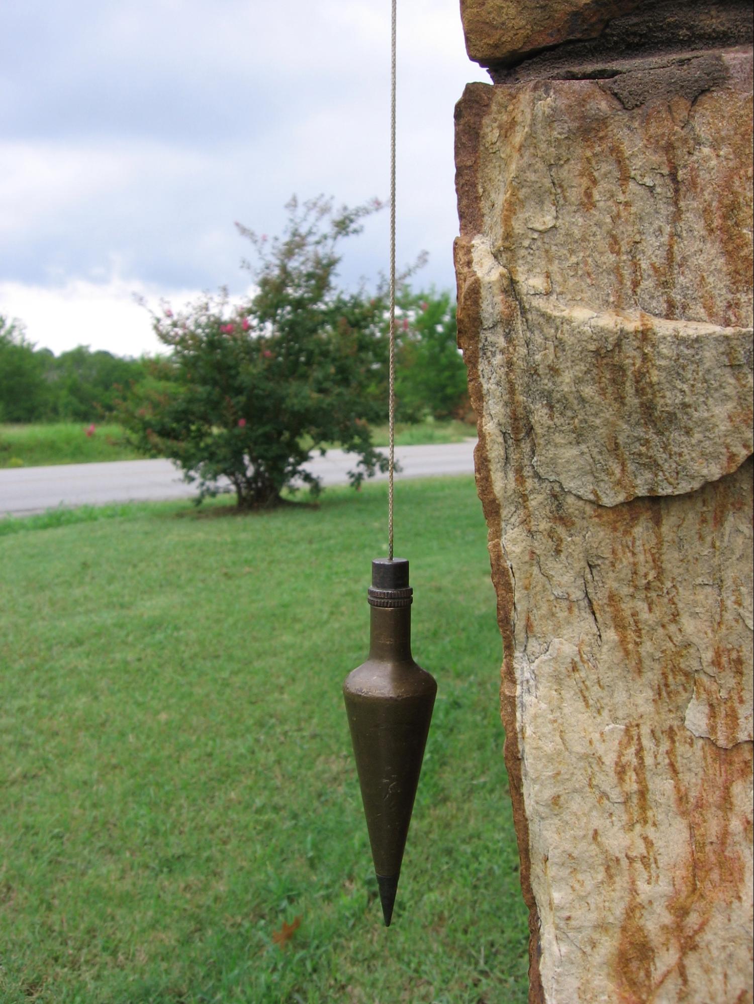 A conical weight suspended by a string next to a crooked sandstone wall.