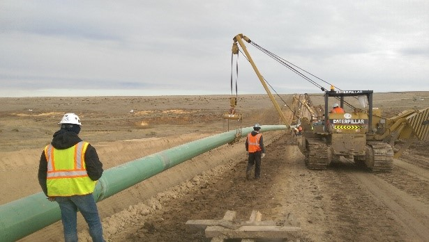 A worker, wearing a safety vest observes from a safe distance as another worker, also wearing a safety vest and hardhat works with an operator of a construction crane to hoist a middle section of a steel pipeline, which is green, so workers seen in the distance can work on the pipeline.