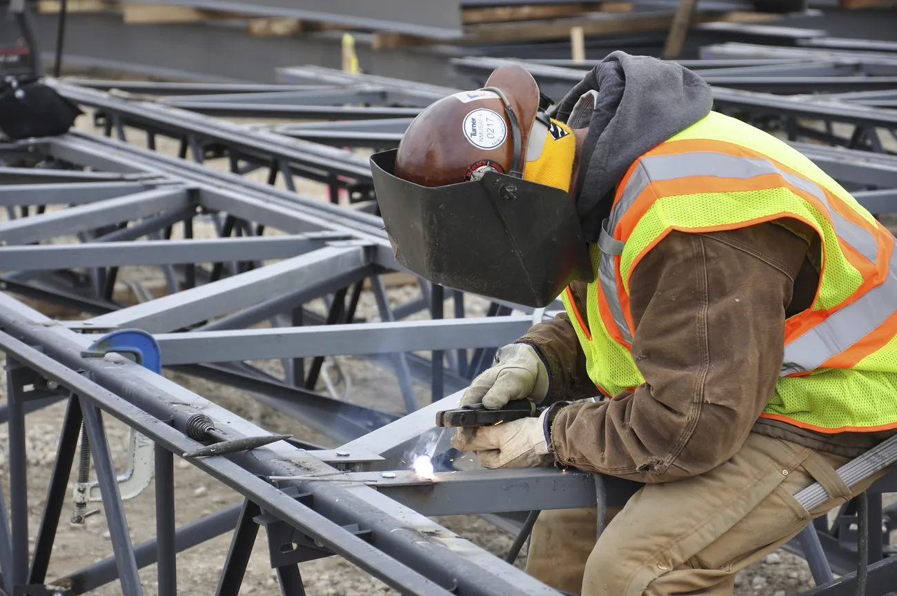 A welder in a high visibility vest with a hard hat-mounted welding hood welds on trusses.