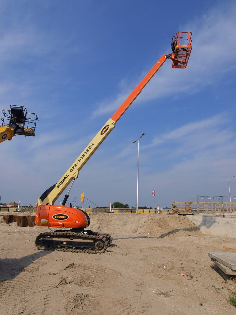 An orange boom lift parked on sand has crawler treads instead of wheels.