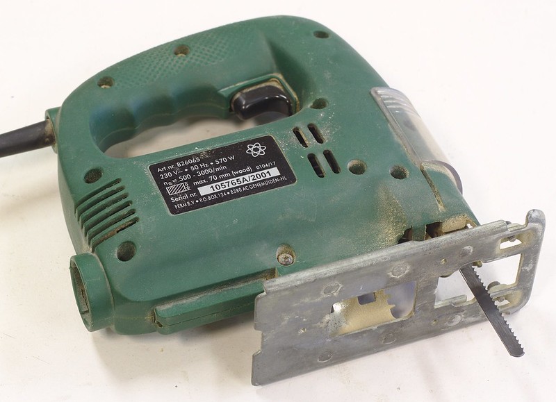 A typical jigsaw. This power tool is green with a small straight metal saw blade affixed to the bottom front of the tool. There is a metal foot at the bottom of the tool through which the saw blade moves up and down when the tool is on.