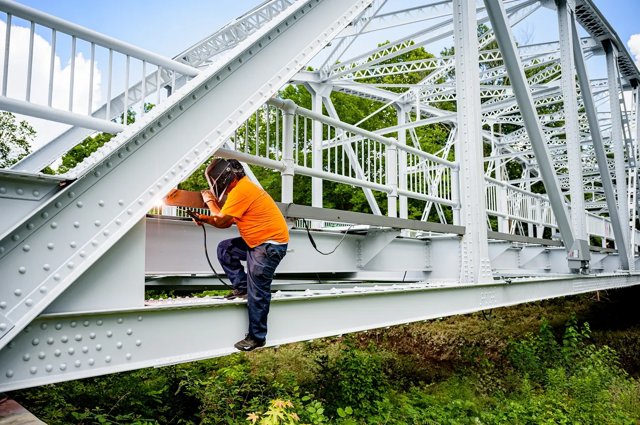 A welder perches on the side of a bridge and welds with one hand, holding onto the bridge with the other hand.