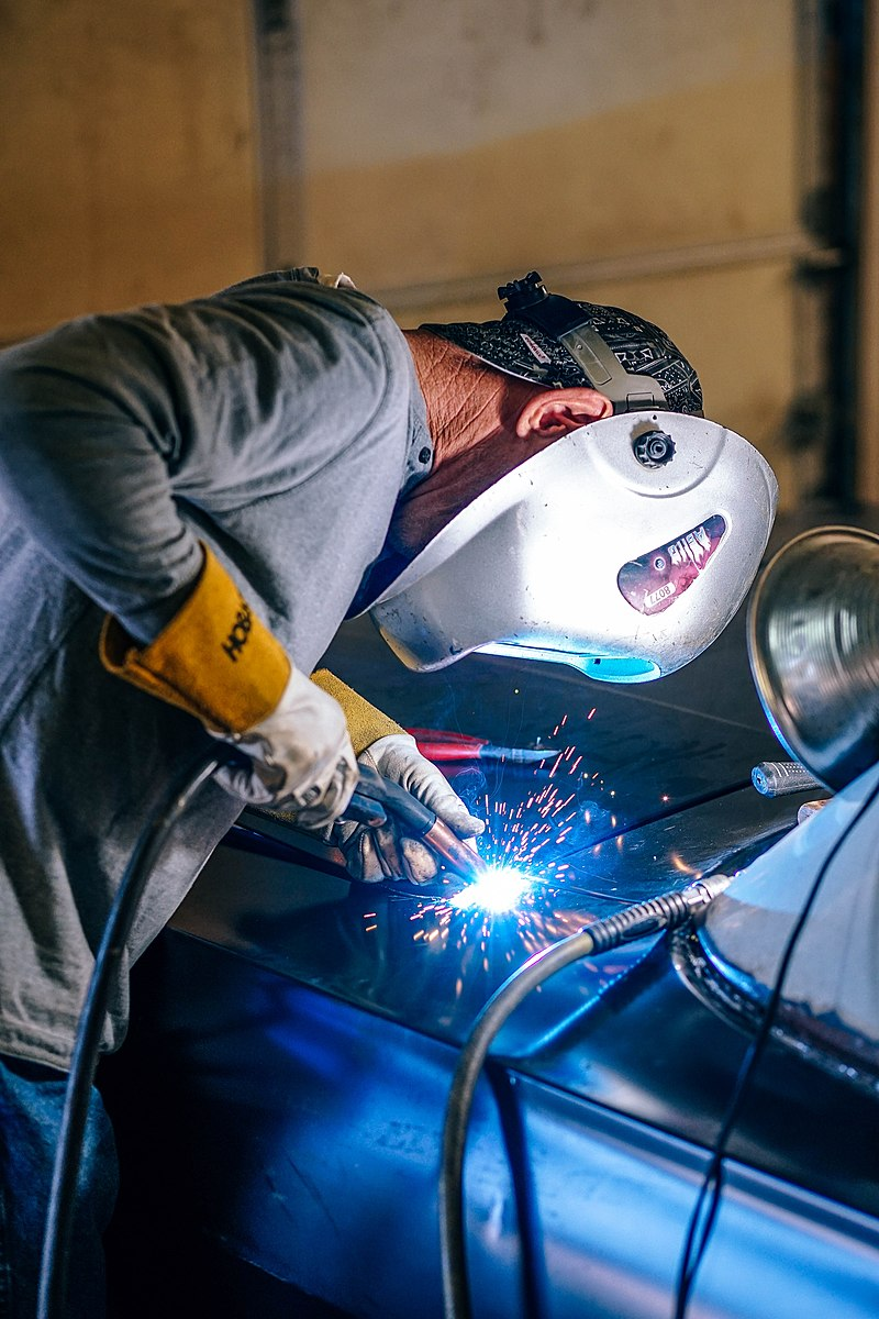 A worker using the GMAW process welds on a vehicle. They are wearing appropriate gauntlet-style gloves, a welders cap, a welding helmet, and a long-sleeved shirt.