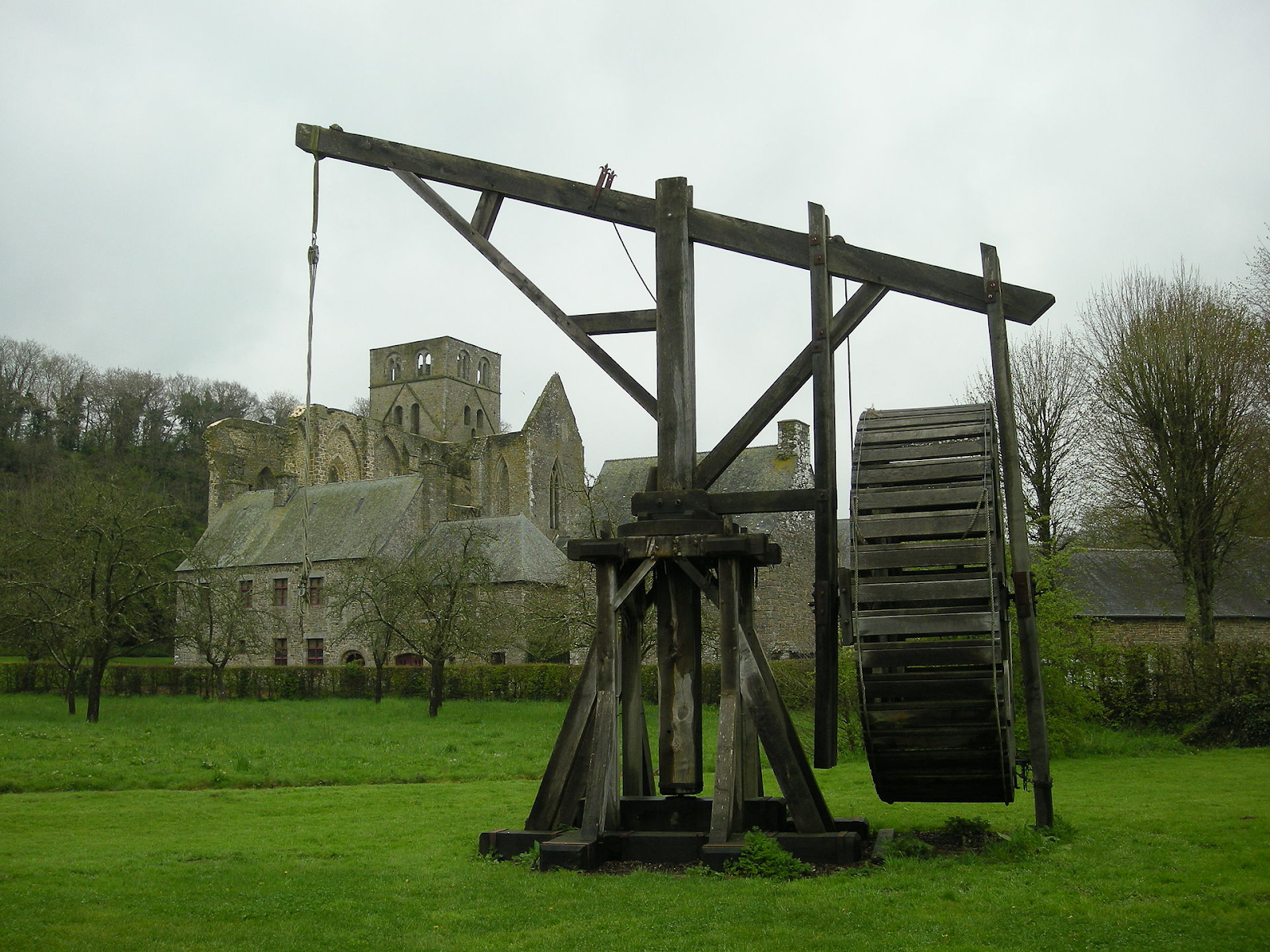 A wooden crane with a large wheel on one side sits in the middle of a field of grass.