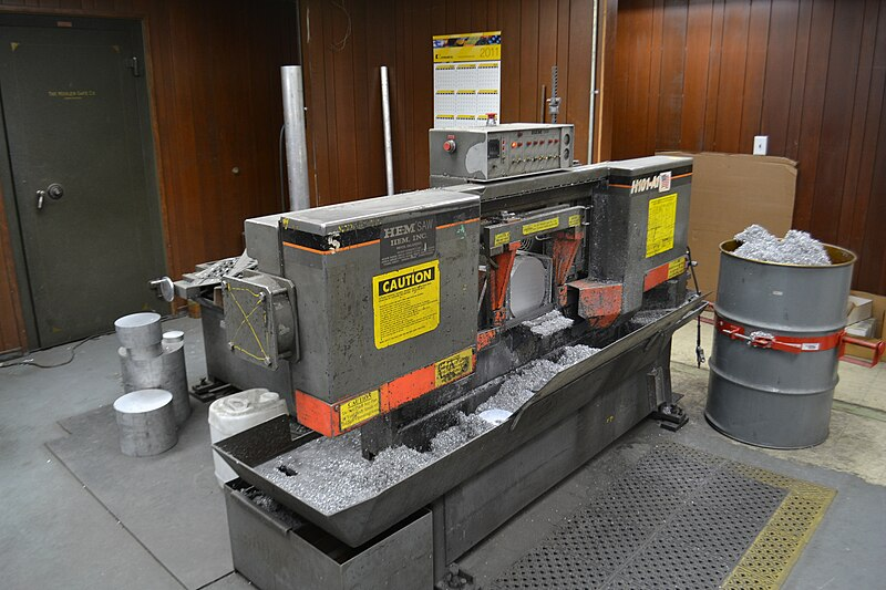 A large gray colored horizontal bandsaw in an industrial shop. The saw is currently stopped and in the down position, and a large aluminum round can be seen still held in the saw’s vice. Several round cutoffs sit on the floor behind the saw and a barrel of aluminum shavings stands to the right.