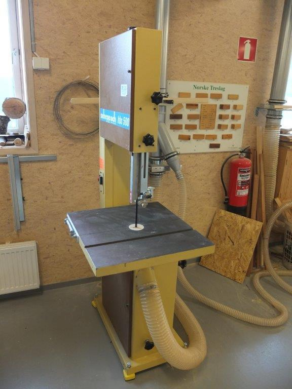 A large upright bandsaw in a wood shop. This band saw is mustard yellow in color with brown panels on the front. It is rectangular, about five or six feet tall and about three feet wide. Midway up the rectangular body, there is a two foot by two foot cutout on the right side. A two foot by two foot table sits at the bottom of the cutout, transverse to the main body. The blade of the saw can be seen coming down from the top part of the body, through a long thin guide, and passing through the center of the table before entering the lower part of the body. In the surrounding shop space can be seen miscellaneous wood remnants and hoses for a dust collection system. Coiled up spare bandsaw blades, a fire extinguisher, and an example of different wood species hang on the wall behind the saw.