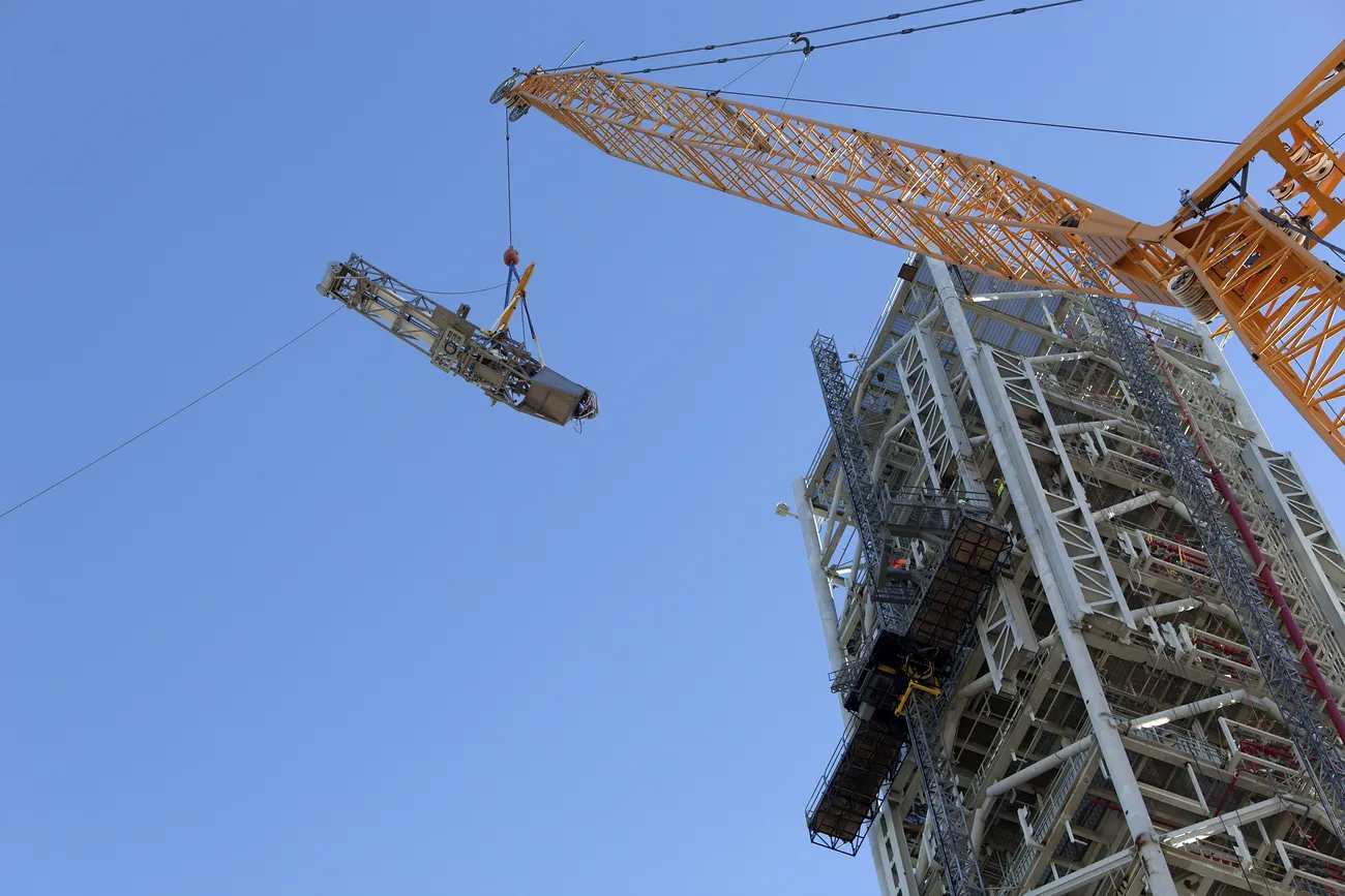 A large tower crane lifts a large load to the building under construction next to it.