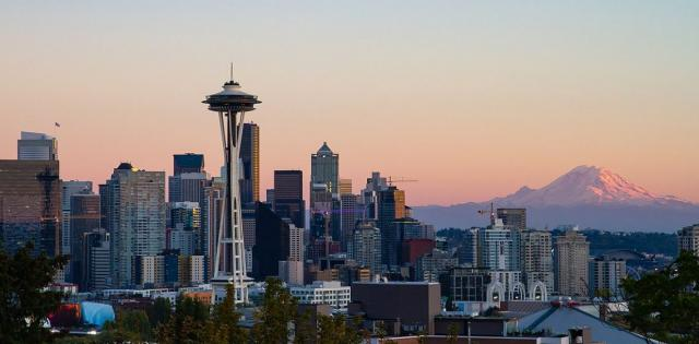 The Seattle city skyline is pictured in the left foreground including tall skyscrapers and the Seattle Space Needle. On the right, Mount Rainier is in the distance. It is sunset and the sky is pink.